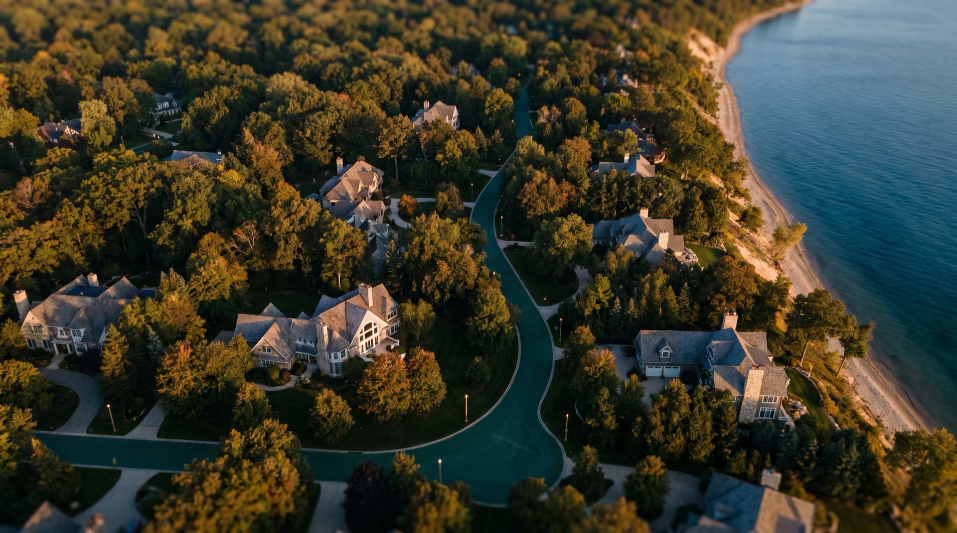 Aerial view over a wooded North Shore lakeside neighborhood, large homes set in tree canopy along the Lake Michigan shoreline at golden hour